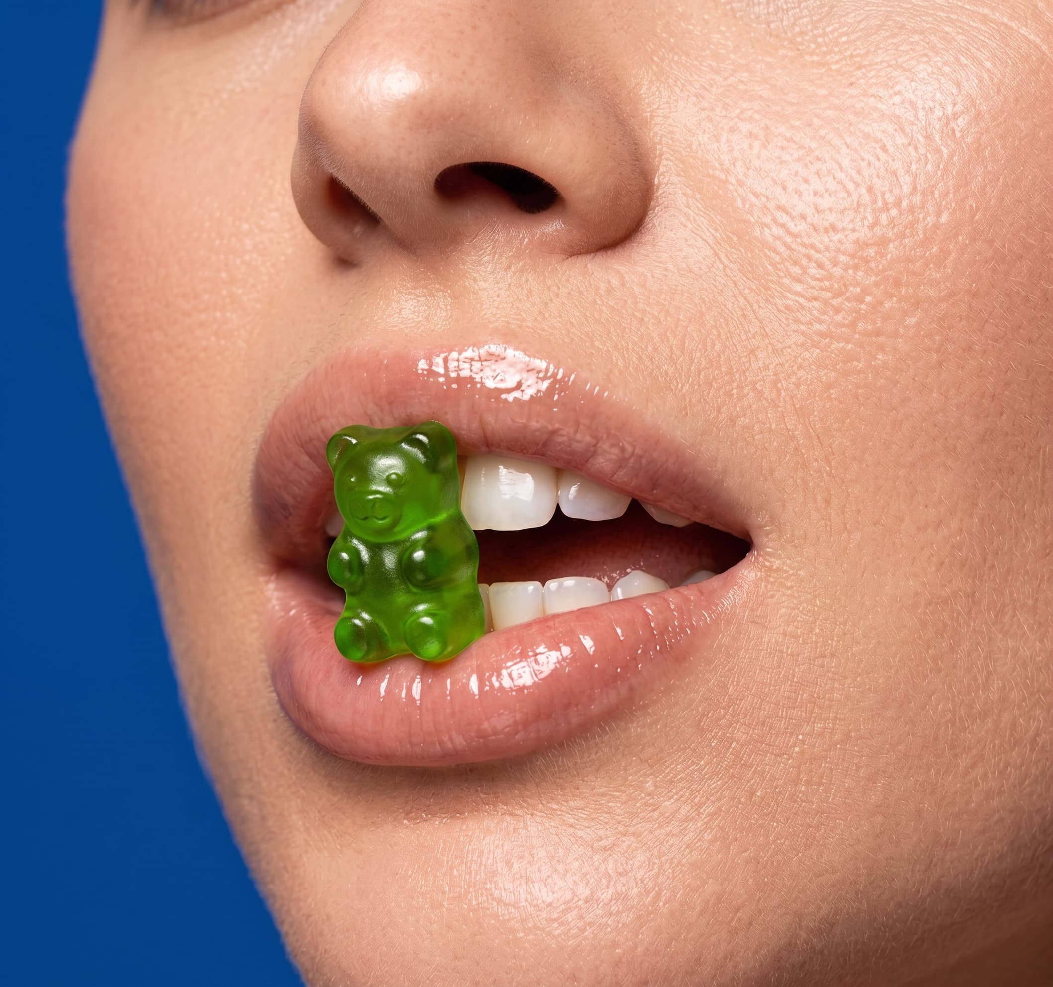 Detalhe de uma gummy verde translúcida junto aos lábios, com fundo azul-cobalto.